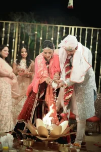 Bride in wine maroon lehenga with pink dupatta and groom in light blue sherwani with pink and white turban performing the havan ritual by pouring rice offerings into a decorative lotus-shaped fire kund during their Hindu wedding ceremony, with smiling family members watching in the background at a destination wedding in India
