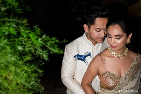 Man in a textured off-white woven bandhgala sherwani featuring a distinctive hand-embroidered blue horse applique on the left chest leaning his head tenderly into the woman beside him who wears a gold and champagne heavily embroidered corset-style saree blouse with a sheer net tissue saree drape a thick gold kundan choker necklace and gold chandelier earrings both with eyes softly closed in a quiet intimate pose with dark lush green bamboo foliage filling the left side of the background at a luxury destination wedding in Rajasthan