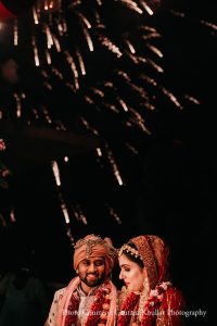 Intimate low-angle portrait of groom in a cream ivory embroidered sherwani with a peach safa turban and layered deep red and maroon jaimala garland looking sideways with a warm smile while the bride beside him in a deep red embroidered bridal lehenga with matching dupatta and gold bridal jewelry looks toward him as cascading white pyrotechnic trails and aerial silver firework bursts explode in the dark night sky filling the upper two-thirds of the frame against a near-black background with a soft red decorative element barely visible at the far upper left at a luxury destination wedding ceremony in Rajasthan