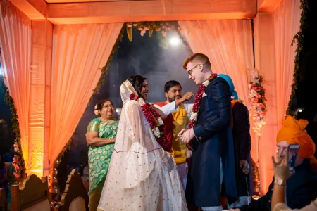 Bride in a white silk lehenga scattered with small gold polka dot embroidery and a matching sheer ivory dupatta with gold border laughing brightly while placing a deep red and white flower jaimala garland around a red-haired groom in a dark navy blue sherwani with brass buttons and black-rimmed glasses who also receives the garland under a draped peach coral silk mandap canopy with floral garlands hanging on the sides as an older woman in a teal green silk saree smiles warmly in the background and white theatrical smoke drifts through the warm orange-lit ceremony space at a luxury destination wedding in Rajasthan