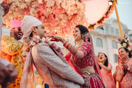 Bride in a heavily embroidered red bridal lehenga with mehndi-covered hands laughing while placing a pink and white rose garland on groom in a cream quilted sherwani with white safa turban as a groomsman in a yellow floral kurta bursts out laughing beside them and female family members clap and smile in the background beneath a massive suspended pink and white floral chandelier at an outdoor destination wedding in Rajasthan