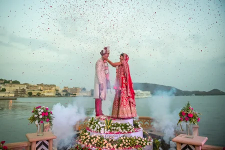 Bride in a red embroidered bridal lehenga and groom in a white floral sherwani with pink dupatta exchanging garlands on an elevated floating stage surrounded by dry ice smoke and a dramatic shower of red and white rose petals with Lake Pichola and the City Palace of Udaipur visible in the background at a luxury destination wedding