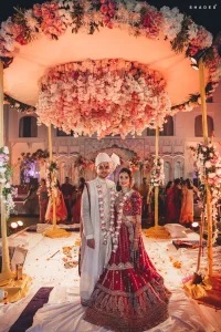 Newlywed groom in a white and cream brocade sherwani with white safa turban wearing a long rose and white flower varmala garland standing beside bride in a heavily embroidered red bridal lehenga with matching flower garland both smiling at the camera under a breathtaking large circular suspended pink and cream floral chandelier mandap with wedding ceremony items visible on the floor and family guests standing in the softly lit heritage palace courtyard behind them at a luxury destination wedding in Rajasthan