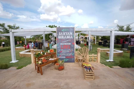 Outdoor daytime pre-wedding event setup with a rustic farm-to-table theme featuring a large chalkboard signboard reading Sanyam Shradha cultivating love since 2021 surrounded by reclaimed wooden furniture wicker peacock chairs and baskets of fresh tomatoes and vegetables under a white tent marquee in an open garden with scrubland hills in the background at a destination wedding in Rajasthan