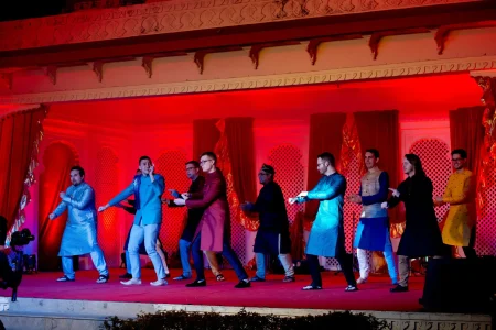 Nine men performing a synchronized choreographed group dance on a wide outdoor stage with a mix of Indian and international guests all dressed in Indian ethnic wear including kurtas in shades of blue teal maroon yellow and black with a few in western trousers under deep red and blue theatrical stage lighting against a backdrop of ornate cream carved heritage palace architecture with decorative jali lattice panels and draped red fabric curtains between arched openings with camera equipment visible at the lower left at a luxury destination wedding sangeet in Rajasthan