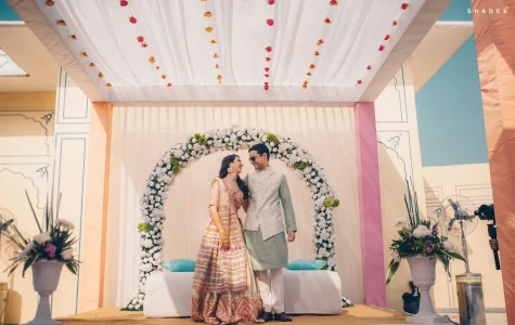 Bride in a vibrant multi-color phulkari mirror work lehenga with ivory dupatta and gold jhumka earrings sharing a laughing moment with groom in a mint green kurta with white embroidered sleeveless jacket and sunglasses as they stand together in front of a full circular white carnation and green flower arch under a white draped fabric tent ceiling with hanging flower drop strings against a pastel blue and peach palace wall at a luxury destination wedding in Rajasthan