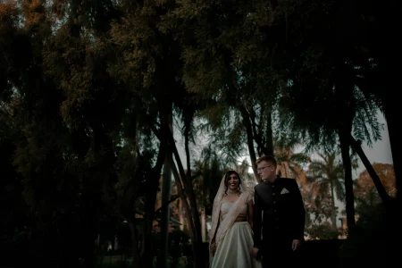 Bride in a white ivory silk saree with a fine gold embroidered lace border dupatta draped over her head and a bold wide silver diamond-set choker necklace smiling softly as she walks beside a red-haired groom in a dark black formal suit with a white pocket square and black-rimmed glasses both photographed from a low dramatic angle beneath a dense canopy of tall dark trees with golden-tipped foliage against a deep charcoal blue dusk sky and a warm terracotta heritage building barely visible in the background at a luxury destination wedding in Rajasthan