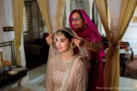 Bride seated in a sunlit palace room during bridal preparation wearing an ivory cream heavily embroidered anarkali suit covered in fine silver and gold zari and sequin work with a champagne dupatta a gold pearl-drop maang tikka and layered gold jhumka earrings her face in soft pink makeup as an older woman in a deep fuchsia magenta embroidered salwar kameez with matching dupatta and glasses carefully drapes and adjusts the bride's dupatta and maang tikka from behind with mehndi-decorated hands and gold bangles with tall cream and gold floor-length curtains ornate white carved arched doorways a Persian rug underfoot and a full-length mirror softly visible in the background at a luxury destination wedding in Rajasthan