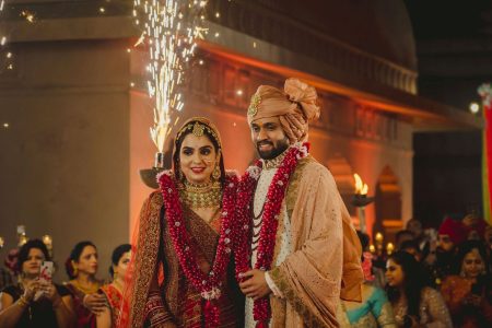 Bride in a deep crimson red heavily embroidered bridal lehenga with a matching red dupatta a large gold polki maang tikka layered gold kundan necklace set and bold red lips smiling softly at the camera while wearing a full deep red and white fresh flower jaimala garland standing beside a bearded groom in a cream white sherwani with a peach rose gold embroidered dupatta draped over his shoulder a peach safa turban with a gold sarpech brooch and his own matching red flower jaimala as silver cold pyrotechnic sparklers burst upward behind them with a sandstone heritage dome structure glowing in warm orange light and a crowd of guests holding phones and celebrating in the blurred background at a luxury destination wedding in Rajasthan