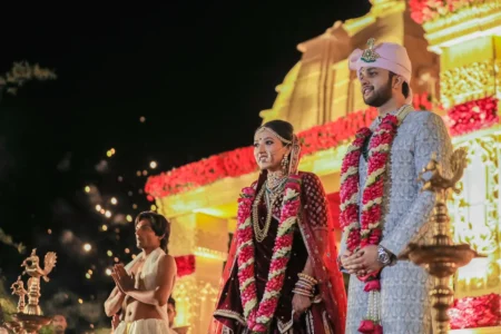 Bride in deep maroon embroidered lehenga with heavy bridal jewellery and a dramatic red and white floral garland, and groom in light blue embroidered sherwani with white turban and emerald necklace, standing before a grand illuminated temple-style wedding mandap decorated with red marigold and yellow floral garlands during a night wedding ceremony at a luxury destination wedding in India