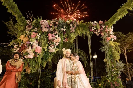 Bride in embroidered pink lehenga and groom in cream sherwani with gold turban seated at their wedding ceremony under an elaborate arch of pink, purple, and white flowers with cascading hanging greens, as fireworks burst in the night sky above at a luxury outdoor destination wedding in Rajasthan