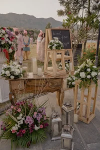 Elegant outdoor wedding welcome table decorated with white pillar candles, silver and wooden lanterns, and fresh mixed floral arrangements in purple, pink, and white, with a chalkboard easel sign and baraat procession visible in the background against Aravalli hills at a destination wedding venue in Rajasthan