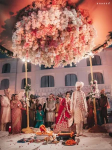 Groom in a white brocade sherwani with white safa turban leading bride in a heavily embroidered red bridal lehenga by the hand as they perform the pheras ritual walk around a sacred havan fire decorated with orange marigold garlands beneath an enormous circular pink peach and white floral chandelier mandap lit with warm amber light while family members in traditional Indian wedding attire watch reverently from the sides with a grand white heritage palace building with arched windows illuminated in the background at a luxury destination wedding in Rajasthan