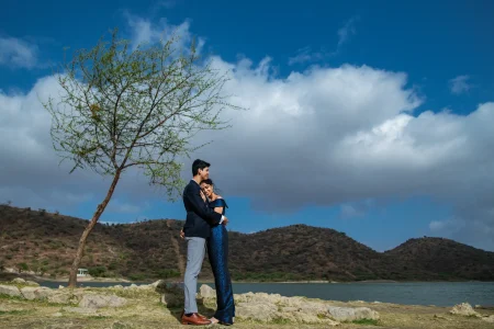 Couple in a close embrace standing near a lone tree on a rocky lakeside with Aravalli hills and blue sky with dramatic white clouds in the background, woman in a royal blue sequin off-shoulder gown resting her head against man in a dark navy suit, during a pre-wedding photoshoot in Udaipur Rajasthan