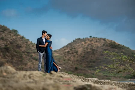 Couple sharing a romantic moment standing on a rocky hilltop overlooking a lake with the Aravalli hills in the background, woman in a royal blue sequin off-shoulder gown and man in a dark navy suit, during a pre-wedding photoshoot at a destination wedding in Udaipur Rajasthan