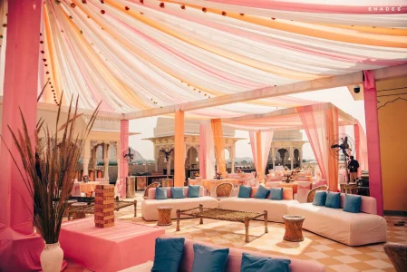 Elegant open-air rooftop wedding event venue decorated with sweeping pink peach and white draped fabric ceiling tent supported by pink poles with tall dried pampas grass in a white vase white low modular sofas with blue and peach cushions wicker chairs bamboo stools and a giant Jenga block set as a games prop with carved Rajasthani sandstone jali arches and a fort-like structure visible on the terrace beyond at a luxury destination wedding venue in Rajasthan