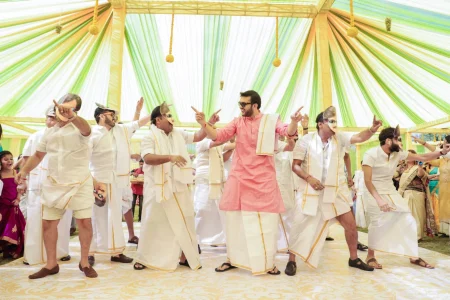 Group of men in traditional white mundu with gold border and face masks performing an energetic synchronized dance performance at a South Indian wedding sangeet, with groom in the center wearing a pink kurta and white mundu dancing under a yellow and lime green draped canopy