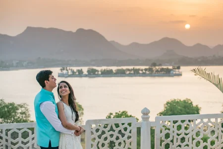 Couple standing close together on a white marble ornate balustrade terrace overlooking Lake Pichola with Jag Mandir Island Palace and Aravalli hills silhouetted against a golden orange sunset sky, man in a turquoise blue nehru jacket and woman in a white lace dress smiling up at each other, during a romantic pre-wedding photoshoot at a luxury palace in Udaipur