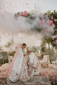 Bride in white floral lehenga and groom in cream sherwani sharing an intimate moment during wedding ceremony rituals as rose petals and pink confetti explode dramatically around them under a lush outdoor floral arch with Aravalli hills in the background at a destination wedding in Rajasthan