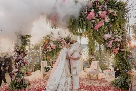 Bride and groom sharing a close romantic moment at the conclusion of their wedding ceremony surrounded by sparklers and pink smoke under an elaborate outdoor arch decorated with pink roses, hydrangeas, and cascading green foliage at a luxury destination wedding in Rajasthan