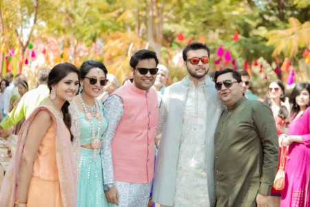 Bride in aqua mint lehenga and groom in silver sherwani posing with three friends in vibrant ethnic outfits against a background of multicolored tassels and pompom hanging decorations at an outdoor mehendi celebration at a destination wedding in India