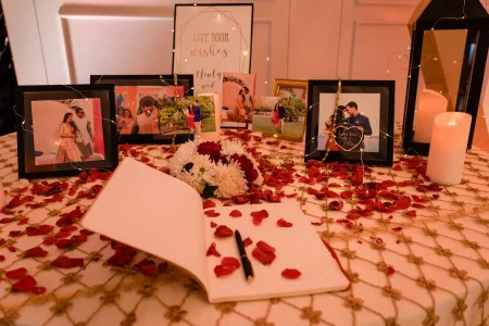 Styled wedding wishing table with an open white guest book and black pen surrounded by scattered red rose petals on a gold embroidered ivory tablecloth with multiple framed pre-wedding couple photographs wrapped in fairy string lights a small white and red flower arrangement white pillar candles in black lanterns and a calligraphy signboard reading Give Your Wishes Newly Wed in the background at a luxury wedding celebration in India