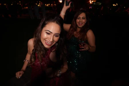 Two young women laughing and dancing with abandon at a dark outdoor wedding cocktail party one wearing a wine red embroidered halter neck dress with curly hair and nose ring and the other in a teal green sequin mini dress holding a drink as green stage lights illuminate the crowded celebration behind them at a luxury destination wedding in India