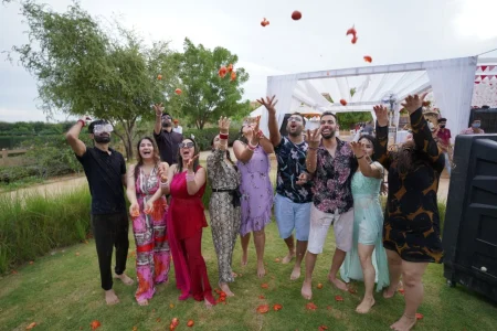 A large group of eight joyful wedding guests in casual colorful outfits throwing tomatoes high in the air simultaneously with arms raised and mouths open in excitement at an outdoor La Tomatina inspired pre-wedding event on a green lawn with a white tent marquee structure and lush monsoon green trees in the background at a destination wedding in Rajasthan