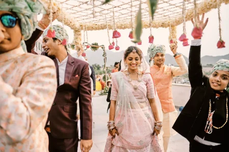 Bride in pink lehenga with heavy gold jewellery walking under white floral hanging canopy during bridal entry with Aravalli hills in backdrop at destination wedding in Rajasthan