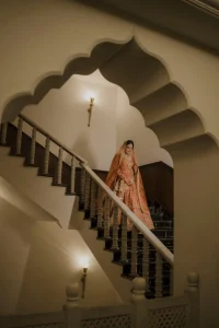 Bride in orange embroidered bridal lehenga descending palace staircase framed by Rajasthani scalloped arch, luxury palace destination wedding in Rajasthan