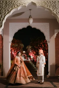 Bride in orange embroidered bridal lehenga posing under Rajasthani Mughal arch with groom in white sherwani and orange safa, palace wedding night portrait, Rajasthan