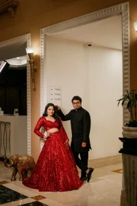 Bride in red sequin lehenga and groom in black sherwani posing in luxury hotel lobby during sangeet night, destination wedding in Rajasthan