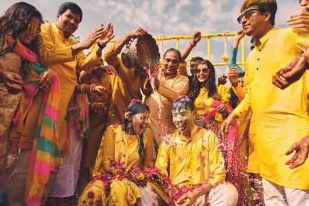 Family pouring water on laughing bride and groom covered in pink rose petals during traditional haldi ritual at destination wedding in Rajasthan