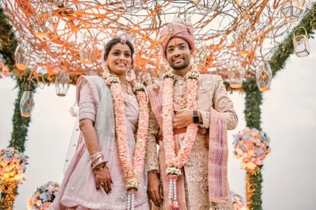 Newlywed bride in blush pink lehenga and groom in gold sherwani wearing varmala posing under organic branch mandap with pastel florals at destination wedding in Rajasthan by Desert Pearl Weddings
