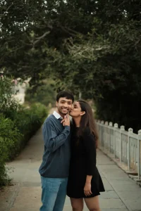 Couple in casual western outfits sharing candid cheek kiss moment along tree-lined garden path during pre-wedding shoot at destination wedding venue in Rajasthan