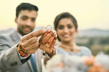 Couple holding up wedding rings on bridal mehndi hands at ring ceremony with mountain hills backdrop at destination wedding in Rajasthan