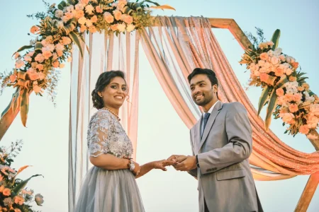 Couple exchanging rings under hexagonal wooden arch with peach shimmer drapery and orange lily floral arrangements at outdoor engagement ceremony at destination wedding in Rajasthan