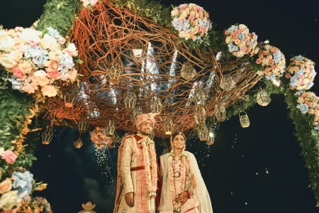 Bride and groom in varmala standing under organic twig mandap canopy with gold geometric lanterns and pastel floral clusters at nighttime destination wedding in Rajasthan