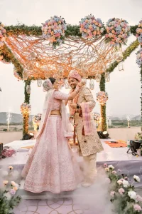 Bride in pink lehenga and groom in gold sherwani exchanging garlands at organic twisted branch mandap with hanging geometric lanterns and pastel florals at dusk destination wedding in Rajasthan