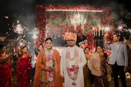 Bride in orange lehenga and groom in white sherwani walking through sparkler aisle in front of red floral wall backdrop with chandelier at luxury destination wedding in Rajasthan